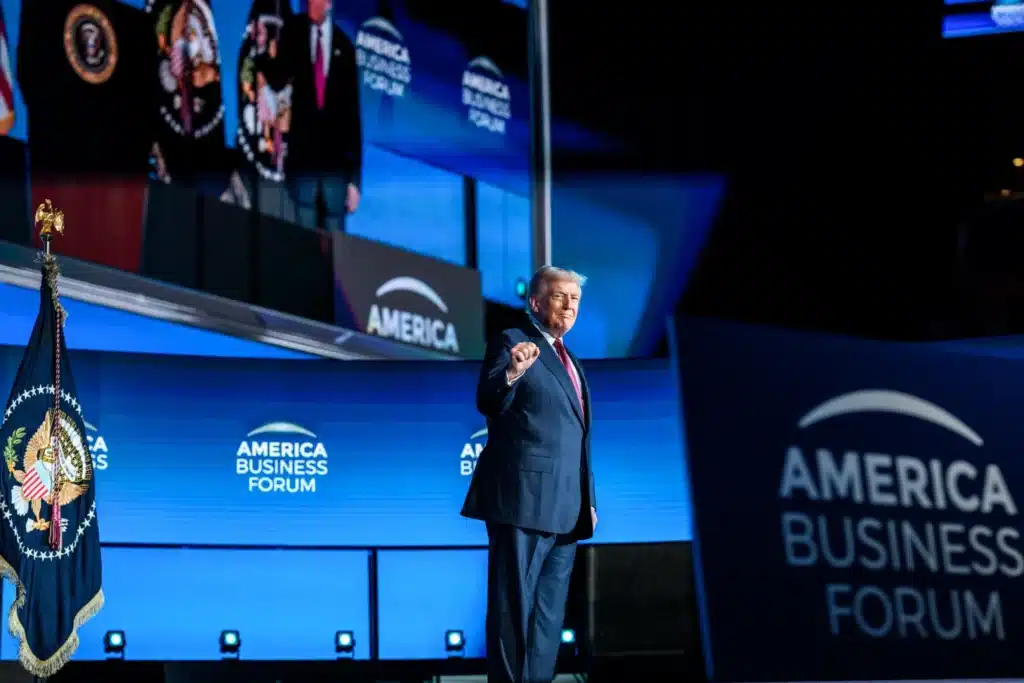 Trump irrumpe en Miami y convierte la alcaldía en una batalla directa contra sus detractores A person stands on stage at the America Business Forum, with a flag in the foreground and large screens