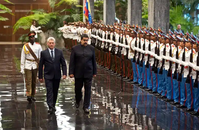 La injerencia del régimen cubano en Venezuela sale a la luz tras captura de Maduro Two men in formal attire walk past a line of uniformed soldiers standing at attention inside a grand hall with tropical plants and flags in the background