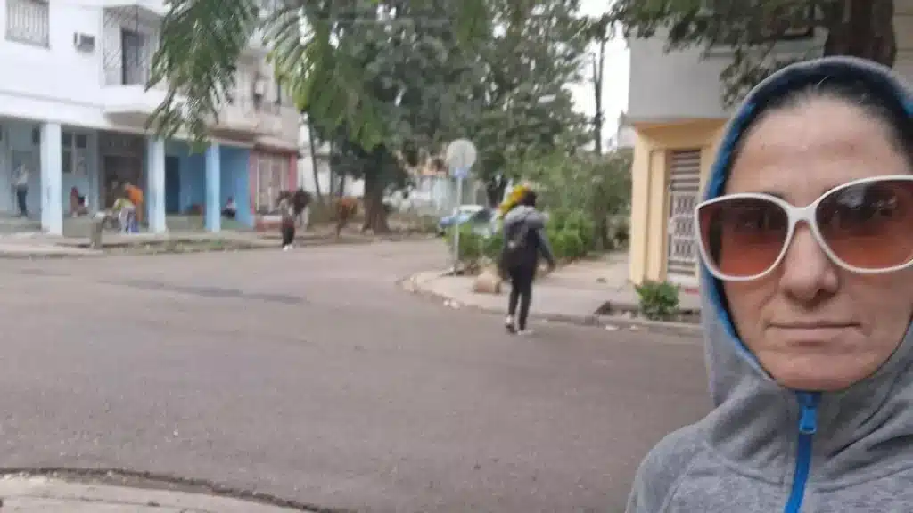 Régimen cubano detiene a Yoani Sánchez y refuerza cerco policial contra opositores en La Habana A person in a gray hoodie and large sunglasses takes a selfie on a quiet, tree-lined urban street. People walk in the background