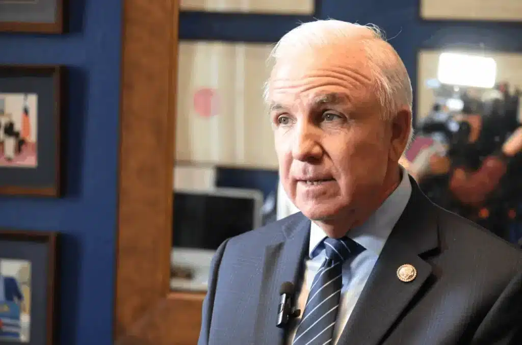A man in a suit and tie speaks in an office. He wears a lapel pin and a microphone. Framed pictures and a camera person are visible in the background.