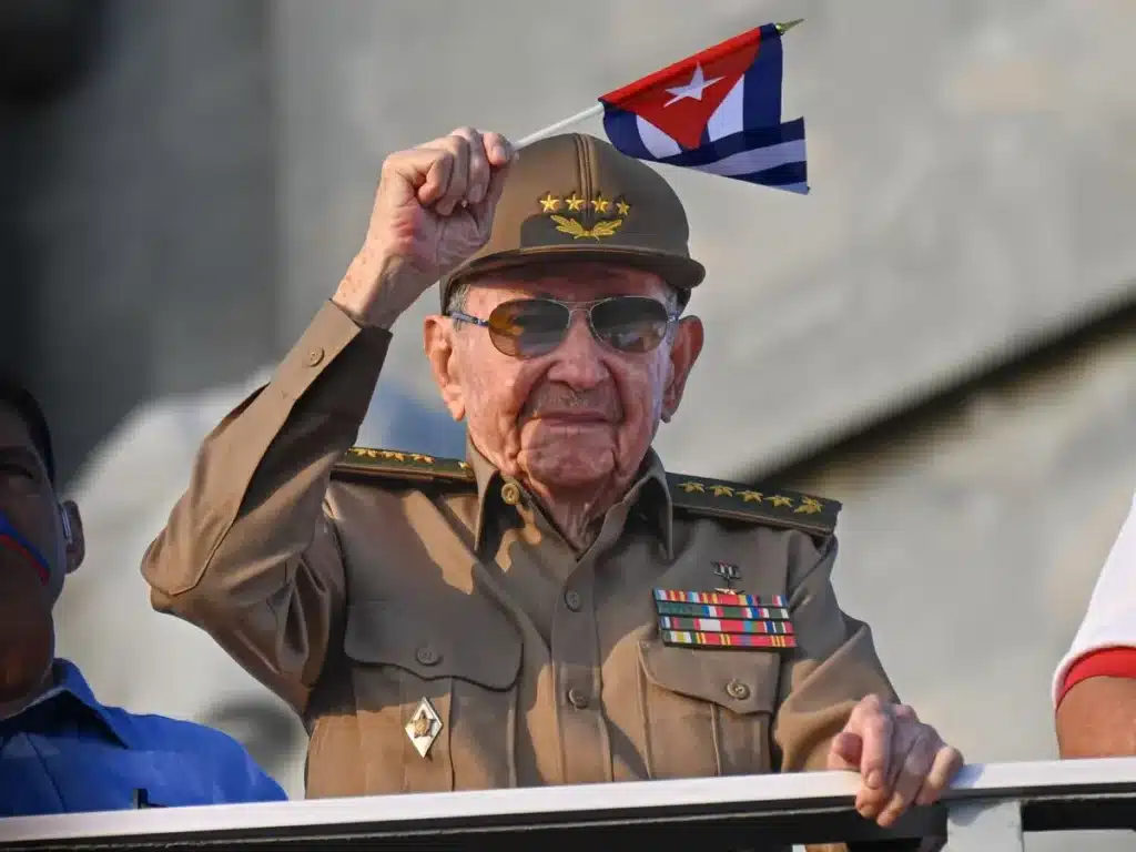 An elderly man in a military uniform and cap salutes with a small Cuban flag. Medals adorn his chest, and he wears sunglasses, exuding a sense of pride.